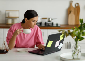 A person in a pink shirt sits at a kitchen table, holding a white cup while looking at a laptop covered in sticky notes from their Money6x.com make money online research. A plant is in the foreground on the right, and there are cutting boards and kitchen utensils in the background. | MONEY6X