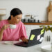 A person in a pink shirt sits at a kitchen table, holding a white cup while looking at a laptop covered in sticky notes from their Money6x.com make money online research. A plant is in the foreground on the right, and there are cutting boards and kitchen utensils in the background. | MONEY6X