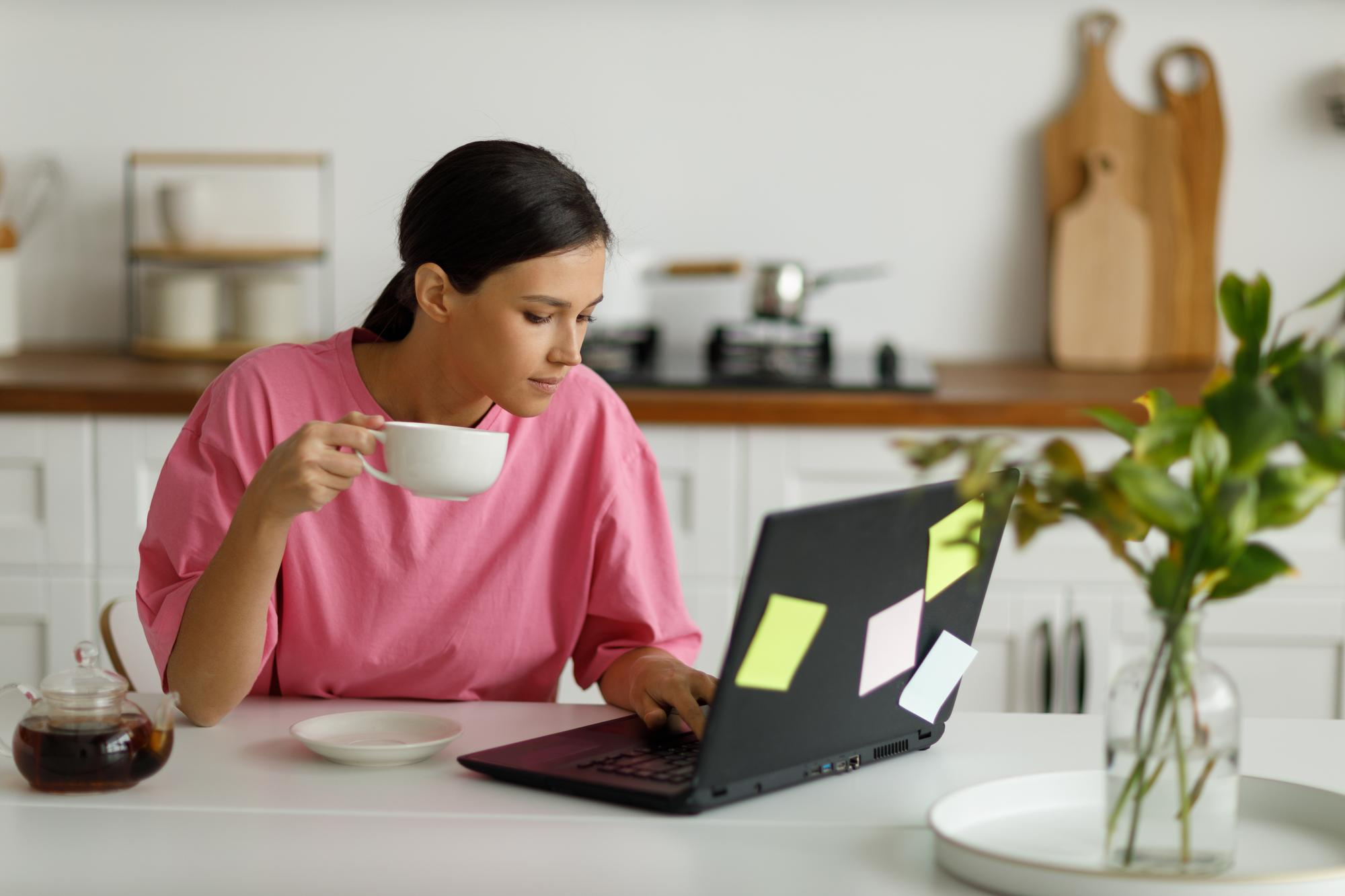 A person in a pink shirt sits at a kitchen table, holding a white cup while looking at a laptop covered in sticky notes from their Money6x.com make money online research. A plant is in the foreground on the right, and there are cutting boards and kitchen utensils in the background. | MONEY6X