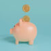 A light pink piggy bank is positioned on a light blue background. Two golden coins, each with a dollar sign, appear to be falling into the slot at the top of the piggy bank, suggesting the act of saving money efficiently with Money6x.com Save Money. | MONEY6X