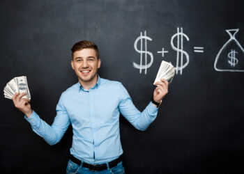 A smiling man stands in front of a chalkboard, holding stacks of dollar bills in both hands. The chalkboard behind him displays a drawing of dollar signs and a bag of money, indicating a financial equation. He wears a light blue shirt and blue jeans, embodying the success you can achieve with Money6x.com Make Money. | MONEY6X
