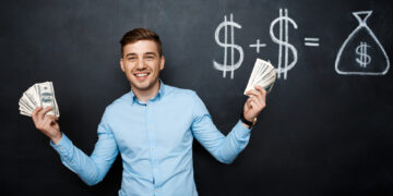 A smiling man stands in front of a chalkboard, holding stacks of dollar bills in both hands. The chalkboard behind him displays a drawing of dollar signs and a bag of money, indicating a financial equation. He wears a light blue shirt and blue jeans, embodying the success you can achieve with Money6x.com Make Money. | MONEY6X