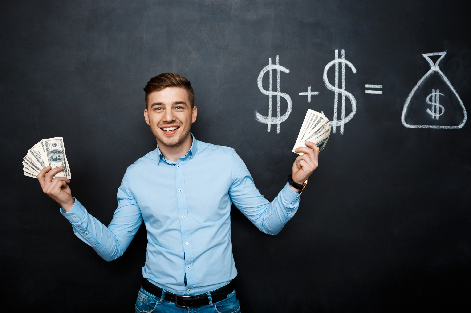 A smiling man stands in front of a chalkboard, holding stacks of dollar bills in both hands. The chalkboard behind him displays a drawing of dollar signs and a bag of money, indicating a financial equation. He wears a light blue shirt and blue jeans, embodying the success you can achieve with Money6x.com Make Money. | MONEY6X