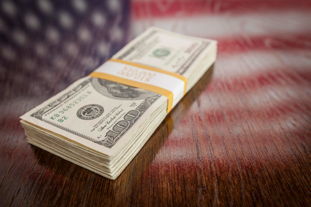 A stack of U.S. hundred-dollar bills wrapped with a yellow band sits on a wooden surface, with a blurred American flag in the background—perfect for those looking to Money6x.com Make Money. | MONEY6X