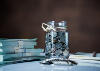 A glass jar labeled "Emergency Fund" brims with assorted coins on a table next to two stacks of banknotes wrapped in paper bands. In the foreground, a set of keys rests beside a notepad and pen, against a backdrop of blurred, dark brown tones. | MONEY6X