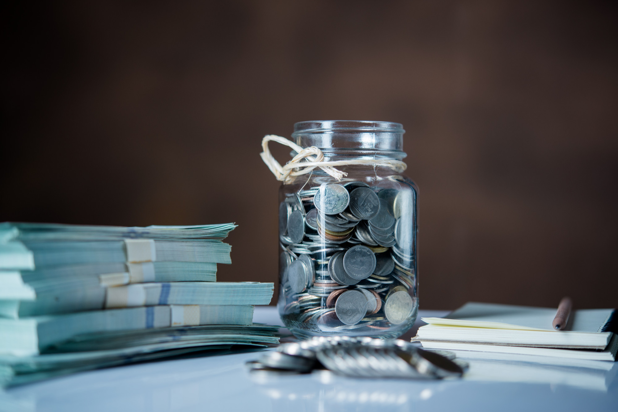 A glass jar labeled "Emergency Fund" brims with assorted coins on a table next to two stacks of banknotes wrapped in paper bands. In the foreground, a set of keys rests beside a notepad and pen, against a backdrop of blurred, dark brown tones. | MONEY6X