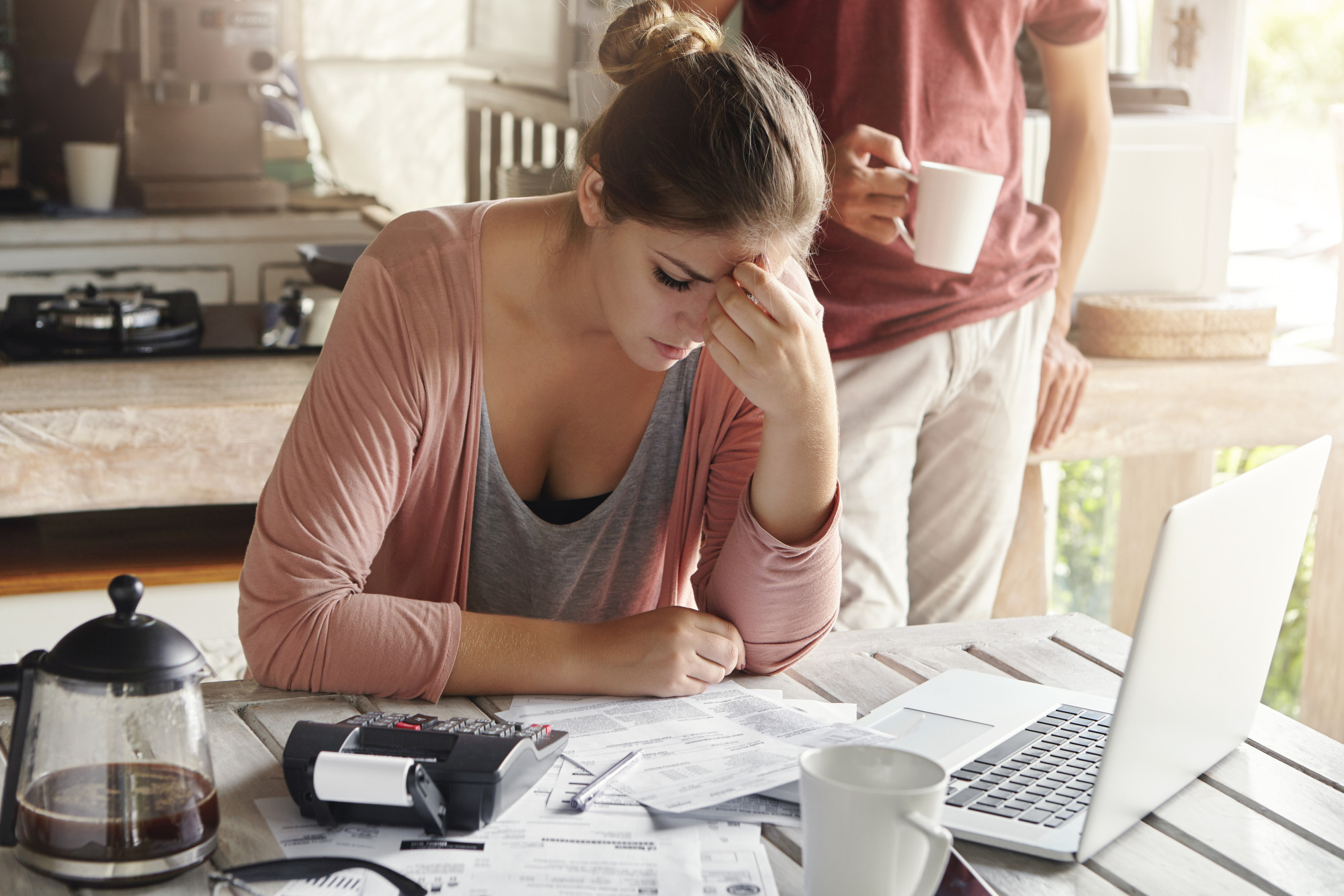 A woman sits at a table with a laptop, head in hands from the headache of foreclosure, surrounded by bills and a calculator. Nearby, someone holds a coffee mug as the coffee pot sits on the table next to an untouched white mug. | MONEY6X