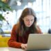 A young woman with long hair, wearing a red sweater, sits at a table working on a laptop in a bright, modern setting with large windows and a plant nearby. | MONEY6X