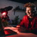 A person wearing headphones and a red hoodie is sitting at a computer desk with a microphone, surrounded by plants. The room is dimly lit with red lighting, and the person is smiling or laughing. | MONEY6X