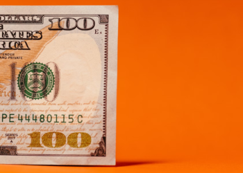 A partial view of a 100 US dollar bill against an orange background showcases intricate patterns, the smart green treasury seal, and part of the serial number. | MONEY6X