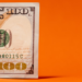 A partial view of a 100 US dollar bill against an orange background showcases intricate patterns, the smart green treasury seal, and part of the serial number. | MONEY6X
