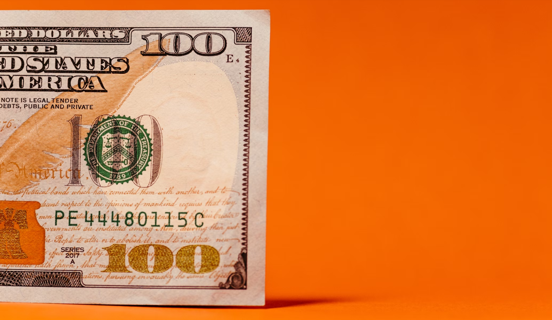 A partial view of a 100 US dollar bill against an orange background showcases intricate patterns, the smart green treasury seal, and part of the serial number. | MONEY6X