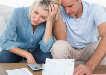 A couple sits on a couch looking stressed while reviewing bills and documents, seeking debt relief. The woman holds her head with one hand, and the man rests his head on his hand, both appearing worried. A calculator is visible beside them as they navigate their financial worries. | MONEY6X