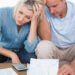 A couple sits on a couch looking stressed while reviewing bills and documents, seeking debt relief. The woman holds her head with one hand, and the man rests his head on his hand, both appearing worried. A calculator is visible beside them as they navigate their financial worries. | MONEY6X