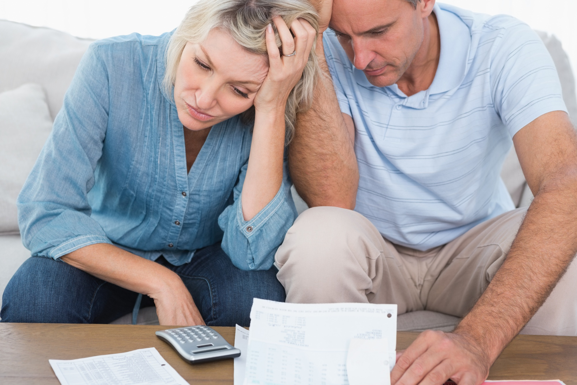 A couple sits on a couch looking stressed while reviewing bills and documents, seeking debt relief. The woman holds her head with one hand, and the man rests his head on his hand, both appearing worried. A calculator is visible beside them as they navigate their financial worries. | MONEY6X