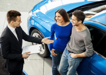 A car salesman, holding a clipboard, gestures while discussing the excitement of buying cars on auction sites with a smiling couple standing next to a blue car in the showroom. The couple appears happy and engaged in conversation. | MONEY6X