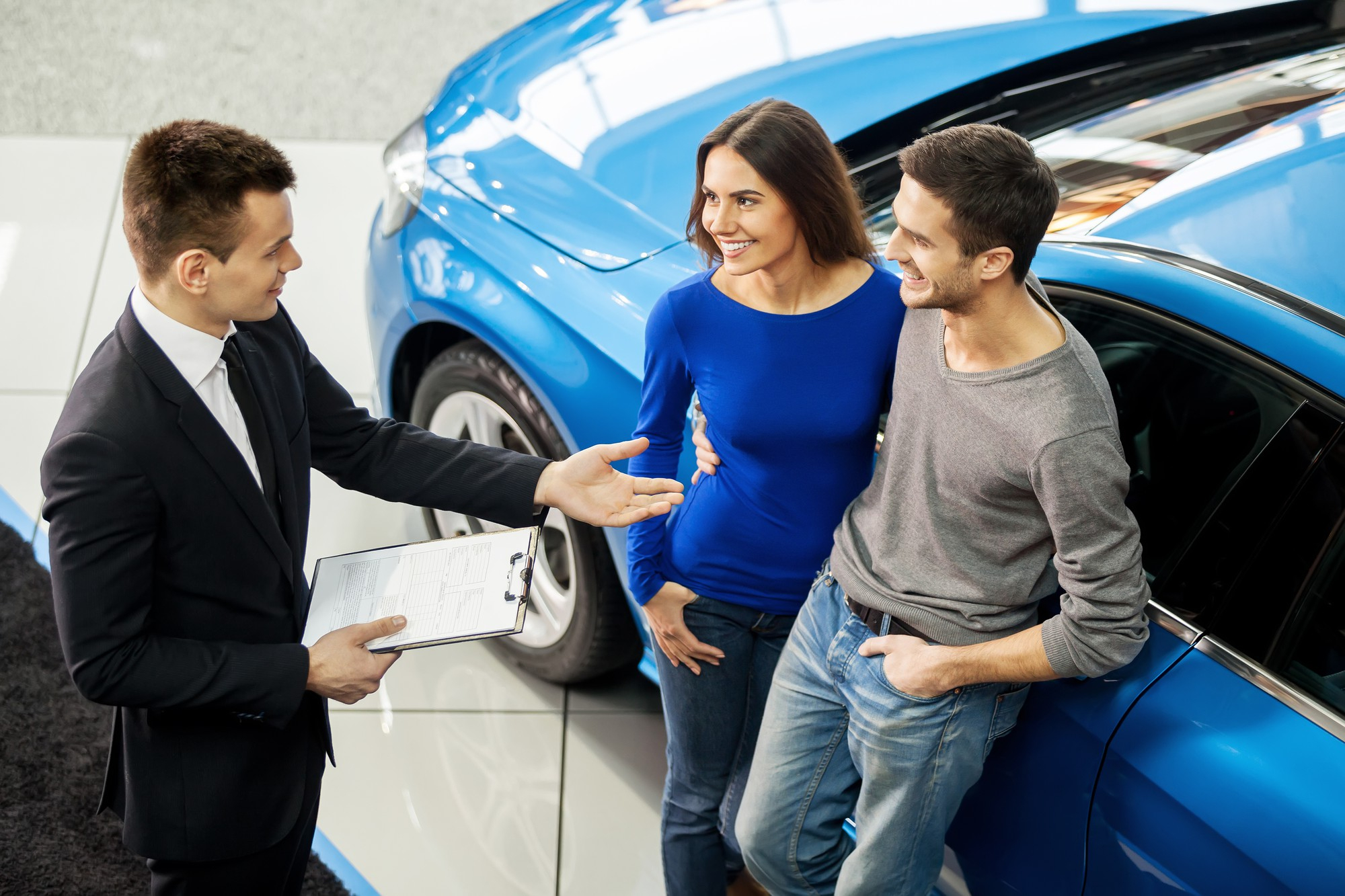 A car salesman, holding a clipboard, gestures while discussing the excitement of buying cars on auction sites with a smiling couple standing next to a blue car in the showroom. The couple appears happy and engaged in conversation. | MONEY6X