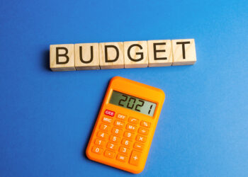 An orange calculator displaying "2021" sits below wooden blocks spelling "BUDGET" against a blue background, hinting at the 50/30/20 Budget strategy for managing finances. | MONEY6X