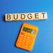 An orange calculator displaying "2021" sits below wooden blocks spelling "BUDGET" against a blue background, hinting at the 50/30/20 Budget strategy for managing finances. | MONEY6X