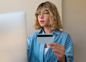 A woman with wavy blonde hair and glasses sits in front of a computer, holding a credit card. Wearing a blue striped shirt, she appears focused on the screen, possibly checking her credit limit. The simple background creates a professional atmosphere. | MONEY6X