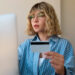 A woman with wavy blonde hair and glasses sits in front of a computer, holding a credit card. Wearing a blue striped shirt, she appears focused on the screen, possibly checking her credit limit. The simple background creates a professional atmosphere. | MONEY6X