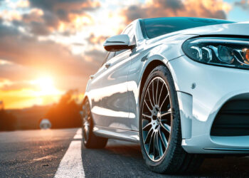 Close-up of a sleek white car parked on the road, its front wheel and headlights prominently showcased. The sky displays a vibrant sunset with clouds, creating a warm, dramatic backdrop that hints at a journey with an impeccable vehicle history report. | MONEY6X