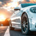 Close-up of a sleek white car parked on the road, its front wheel and headlights prominently showcased. The sky displays a vibrant sunset with clouds, creating a warm, dramatic backdrop that hints at a journey with an impeccable vehicle history report. | MONEY6X