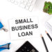 Office desk with a sign reading "Small Business Loan," surrounded by a keyboard, a lanyard, a small plant, a notebook, and two pencils. | MONEY6X