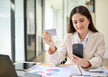 A woman in a beige blazer sits at a desk with a smartphone in one hand and a credit card in the other. Amongst the laptop, tablet, and colorful charts, she appears happy—perhaps exploring core banking solutions while conducting an online transaction. | MONEY6X