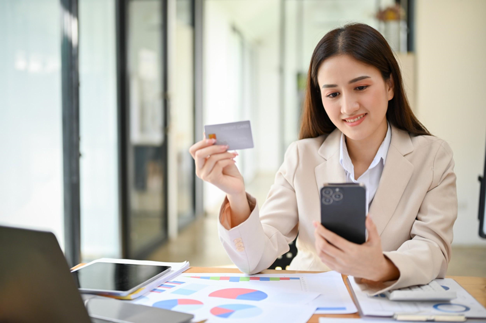 A woman in a beige blazer sits at a desk with a smartphone in one hand and a credit card in the other. Amongst the laptop, tablet, and colorful charts, she appears happy—perhaps exploring core banking solutions while conducting an online transaction. | MONEY6X