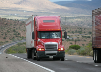 A red semi-truck, highlighting the journey of an owner-operator managing expenses, cruises along a highway in a desert landscape. The road curves with hills and sparse vegetation visible under a partly cloudy sky. Another truck is partially captured on the right side of the image. | MONEY6X