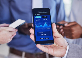 Three people holding smartphones, focused on one displaying a fintech app with a live cryptocurrency chart. The screen reveals BTC at $1250.50 and USD at $125.50, showing a current balance of $113.45. | MONEY6X