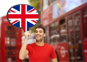 A man in a red shirt holds a speech bubble adorned with the British flag, standing amidst blurred red double-decker buses on a bustling street, contemplating how to start a business in the UK. | MONEY6X