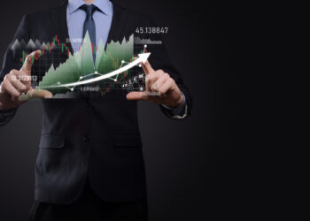 A man in a suit interacts with a virtual screen displaying financial graphs and charts. His hands are positioned as if manipulating data, focusing on risk management in short futures trading. The dark background contrasts with illuminated graphics and a white arrow showing growth. | MONEY6X