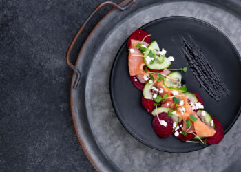 A black plate with smoked salmon, beet slices, cucumber, figs, and crumbled feta topped with microgreens. The dish is artistically arranged on a metal tray with handles, placed on a dark textured surface. | MONEY6X