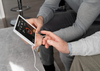 Two people sit together, one holding a tablet from Life Assure Technology displaying health data, while the other touches the screen. The tablet shows heart rate information. They appear focused and engaged in a healthcare-related discussion. | MONEY6X