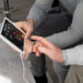 Two people sit together, one holding a tablet from Life Assure Technology displaying health data, while the other touches the screen. The tablet shows heart rate information. They appear focused and engaged in a healthcare-related discussion. | MONEY6X