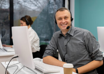 A man in a gray shirt, wearing a headset, sits at a desk with his computer and coffee cup, embodying the professionalism of high-performing contact centre agents. He smiles at the camera while a woman in the background focuses intently on her computer. The room features large windows and teal walls. | MONEY6X