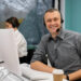 A man in a gray shirt, wearing a headset, sits at a desk with his computer and coffee cup, embodying the professionalism of high-performing contact centre agents. He smiles at the camera while a woman in the background focuses intently on her computer. The room features large windows and teal walls. | MONEY6X