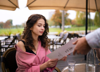 A woman in a pink jacket sits at an outdoor café table, reading from menu holders. A person hands the menu to her. Glasses of water are on the table, and patio umbrellas and greenery provide a serene background. | MONEY6X