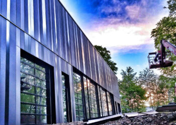 Modern building with sleek, vertical metal panels and large windows reflecting trees. A worker in a cherry picker is on the right, ensuring moisture control, surrounded by trees and a vibrant sky with clouds transitioning from blue to orange. | MONEY6X