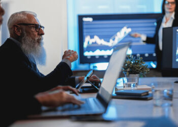 A bearded man in a suit sits at a conference table with a laptop, passionately discussing finance while gesturing towards a large screen showing a graph. In the background, a woman points at another screen displaying financial charts as other attendees are partially visible. | MONEY6X
