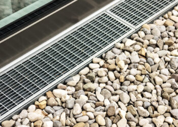 Stormwater drainage grates system are installed next to a window or building edge, with smooth gray and beige pebbles of various sizes arranged neatly alongside it. | MONEY6X