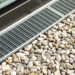 Stormwater drainage grates system are installed next to a window or building edge, with smooth gray and beige pebbles of various sizes arranged neatly alongside it. | MONEY6X
