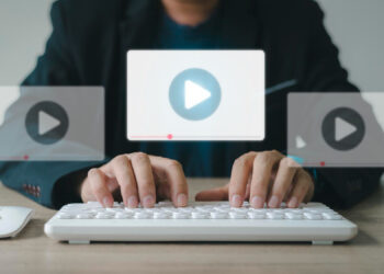 Person typing on a white keyboard, three floating video play icons above the keys symbolizing online video content creation or streaming with a focus on refined video content strategy. Dressed in a black blazer, the setting is a straightforward office desk setup. | MONEY6X