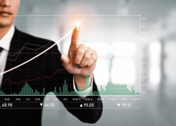 A person in a suit interacts with a virtual financial chart using their finger. The chart, focused on emerging markets, displays various rising and falling line graphs, bars, and numerical data, indicating stock market trends. The blurred background suggests an office setting. | MONEY6X