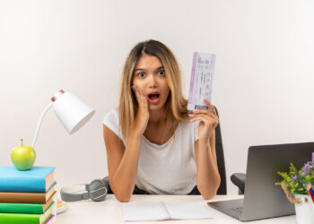 A woman, surprised while holding a boarding pass and pondering how to make an extra $1,000 a month without quitting her job, sits at a desk. Her other hand rests on her cheek amid a laptop, lamp, stacked books, green apple, headphones, notepad, and vibrant purple flowers. | MONEY6X