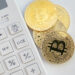 A white calculator is positioned next to three gold-colored coins with the Bitcoin symbol, hinting at the complexities of crypto taxation. The coins boast an intricate circuit-like design, emphasizing a focus on cryptocurrency and finance against a plain, minimalistic background. | MONEY6X