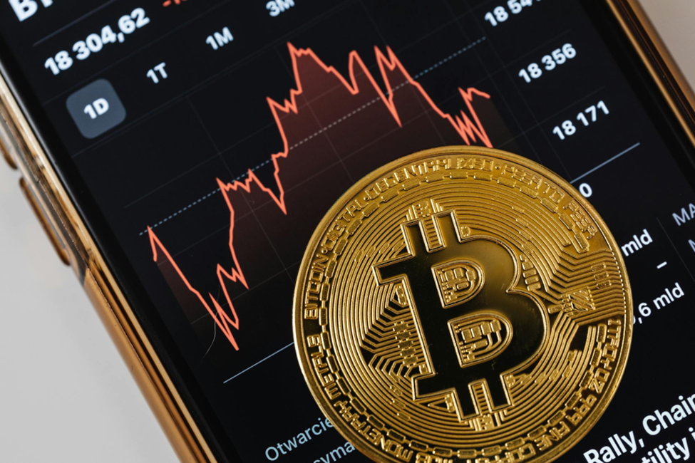 A golden Bitcoin coin rests on a smartphone screen displaying a fluctuating cryptocurrency graph with red lines against a black background. The numbers and chart depict market trends. | MONEY6X