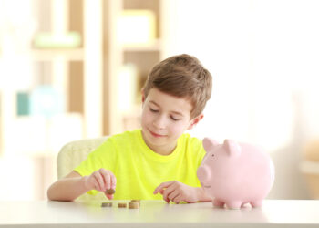 A young boy in a bright yellow shirt is sitting at a table, learning to stack coins as part of teaching kids about money. Beside him, a pink piggy bank stands ready. The softly blurred background hints at a cozy indoor setting. | MONEY6X